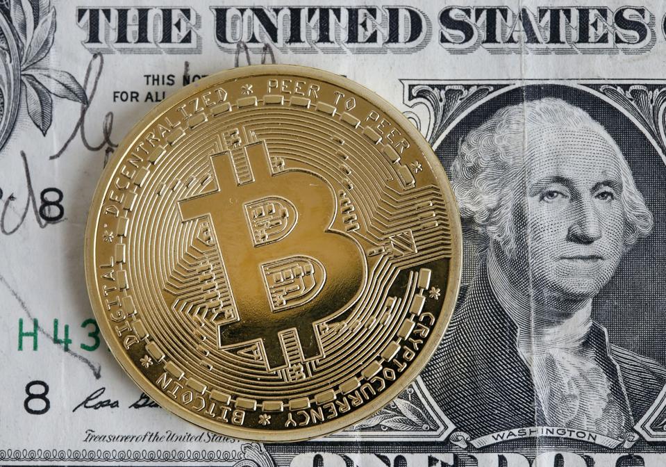 A metal bitcoin sitting atop a paper U.S. dollar bill.